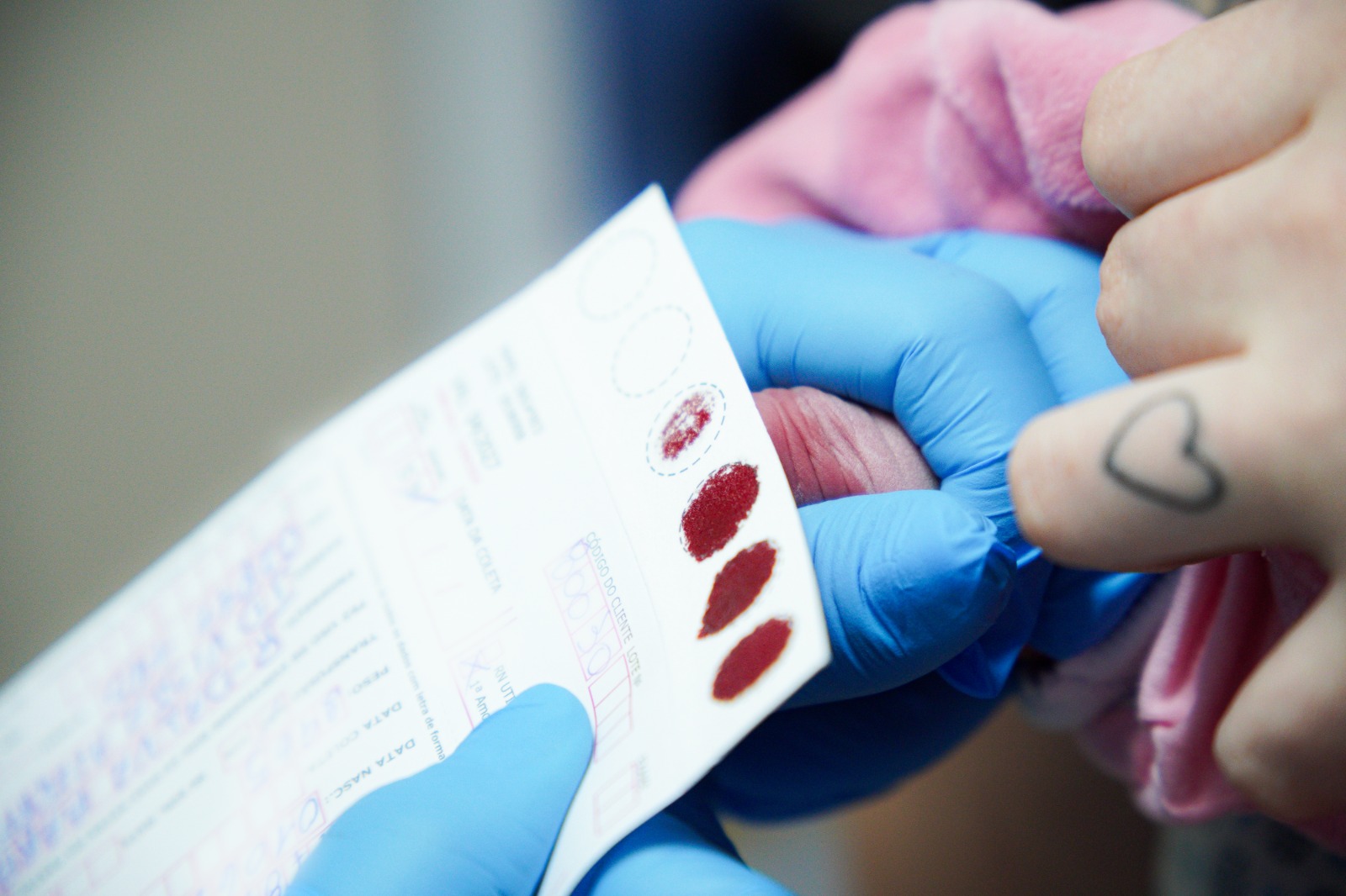 A imagem mostra um close-up de um procedimento de coleta de sangue por punção digital para realização de teste rápido ou exame laboratorial.  Em destaque, uma profissional utilizando luvas descartáveis azuis segura cuidadosamente o dedo de uma pessoa enquanto coleta pequenas gotas de sangue em um cartão ou formulário específico para análise. Sobre o cartão, é possível observar várias gotas de sangue depositadas em áreas circulares demarcadas para a amostra.  A pessoa atendida veste uma peça de roupa rosa e tem uma pequena tatuagem em formato de coração em um dos dedos visíveis. O enquadramento aproxima a cena dos detalhes das mãos e do material de coleta, enquanto o fundo aparece desfocado, direcionando a atenção para o procedimento.  A fotografia transmite um ambiente de cuidado e atendimento em saúde, ilustrando uma etapa comum de testagem rápida ou exames que utilizam amostras de sangue obtidas por punção na ponta do dedo.