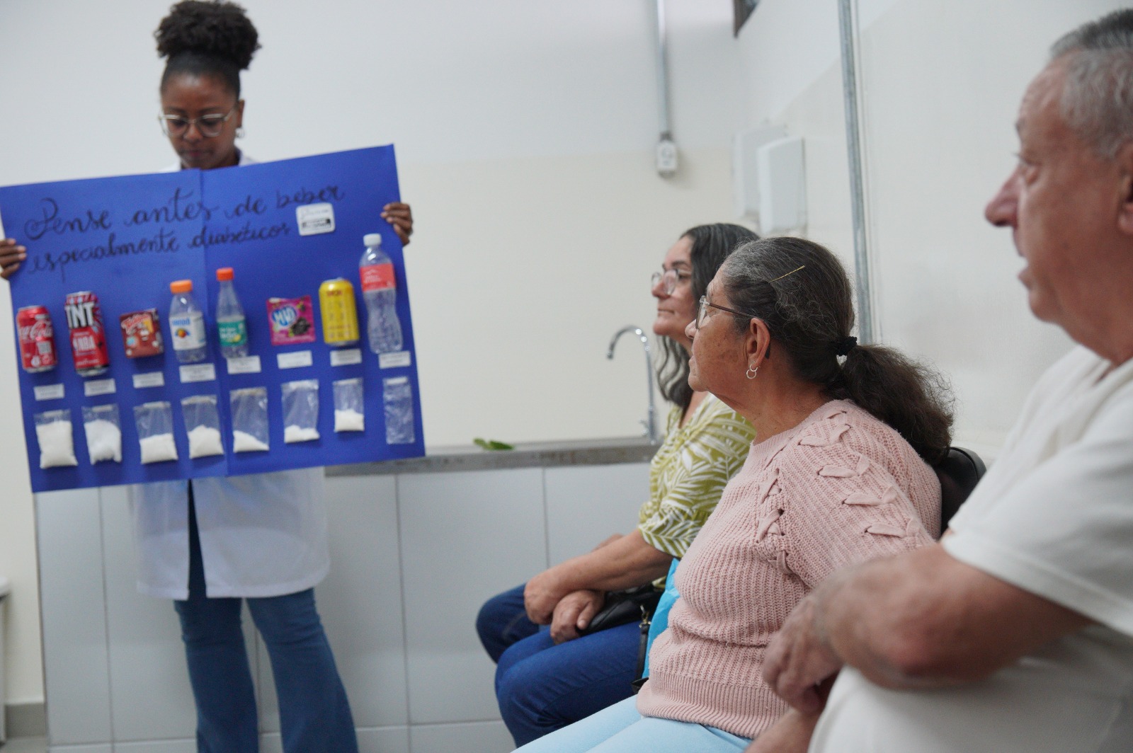 A imagem mostra uma atividade educativa em um ambiente de saúde, provavelmente uma unidade básica. Uma profissional, usando jaleco, está em pé segurando um cartaz azul com a frase “Pense antes de beber, especialmente diabéticos”. No cartaz, há exemplos de bebidas (como refrigerantes, sucos e outras) acompanhadas de saquinhos com a quantidade de açúcar correspondente a cada uma, de forma visual.  Sentados à frente dela, três adultos — dois idosos e uma mulher de meia-idade — observam atentamente a explicação. O ambiente é simples, com pia e parede branca ao fundo, sugerindo uma sala de atendimento ou educação em saúde.