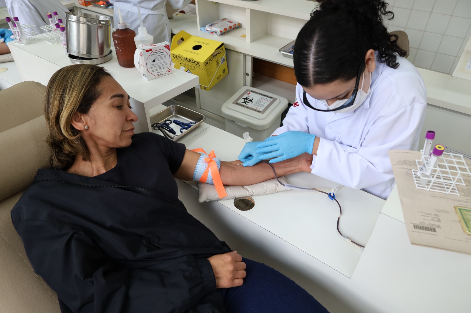 A foto mostra uma profissional de saúde realizando uma coleta de sangue em uma mulher sentada em uma cadeira reclinável, em um ambiente clínico ou laboratório. A paciente está com o braço esquerdo apoiado sobre uma almofada branca e utiliza um garrote azul preso por um laço laranja acima do cotovelo para facilitar o procedimento. Ela veste uma camiseta preta e um casaco escuro, mantendo uma expressão tranquila enquanto observa a coleta. A profissional de saúde usa jaleco branco, máscara de proteção, óculos de proteção apoiados sobre o rosto e luvas descartáveis azuis. Ela está concentrada na punção venosa, segurando a agulha e o equipamento de coleta com cuidado. Ao redor da bancada há diversos materiais utilizados em procedimentos laboratoriais, como suportes com tubos de coleta de sangue de tampas roxas, bandejas metálicas com instrumentos, frascos de antissépticos, recipientes para descarte de materiais perfurocortantes e uma caixa para descarte de resíduos.