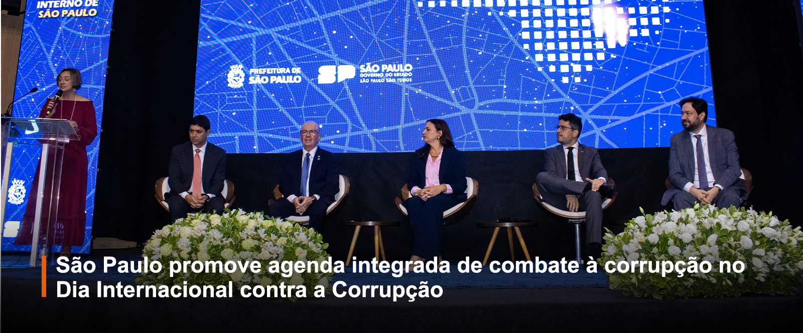 Cinco pessoas sentadas enfileiradas em frente a um telão azul enquanto uma mulher em pé fala ao microfone