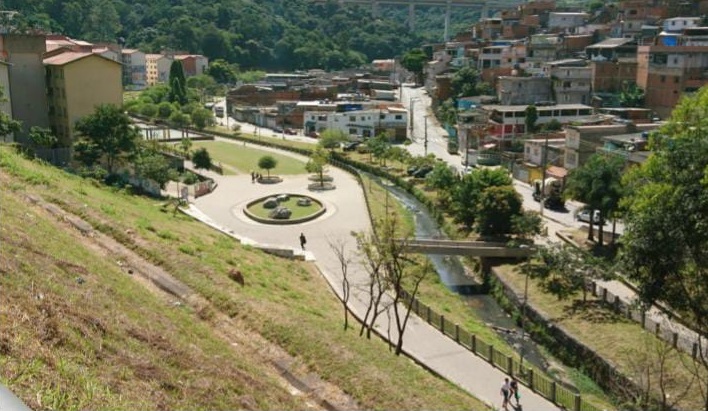 A imagem mostra uma área urbana vista de um ponto mais alto, como uma encosta ou morro. À esquerda, há uma ladeira gramada e inclinada. No centro da imagem, há uma pequena praça com um caminho circular de concreto, algumas árvores plantadas ao redor e um espaço gramado no meio. A praça é cercada por uma ciclovia ou pista de caminhada que segue ao lado de um pequeno córrego com água, que é atravessado por uma ponte estreita de pedestres.  À direita da praça e do córrego, há muitas casas e prédios residenciais construídos de forma bem densa, característica de uma comunidade urbana. As casas são simples, algumas de tijolo exposto, e estão próximas umas das outras. Ao fundo, há uma floresta densa com muitas árvores e uma ponte de rodovia elevada cruzando as montanhas ao longe. O céu está limpo, sugerindo um dia ensolarado. Algumas pessoas aparecem caminhando nas trilhas e calçadas da área.