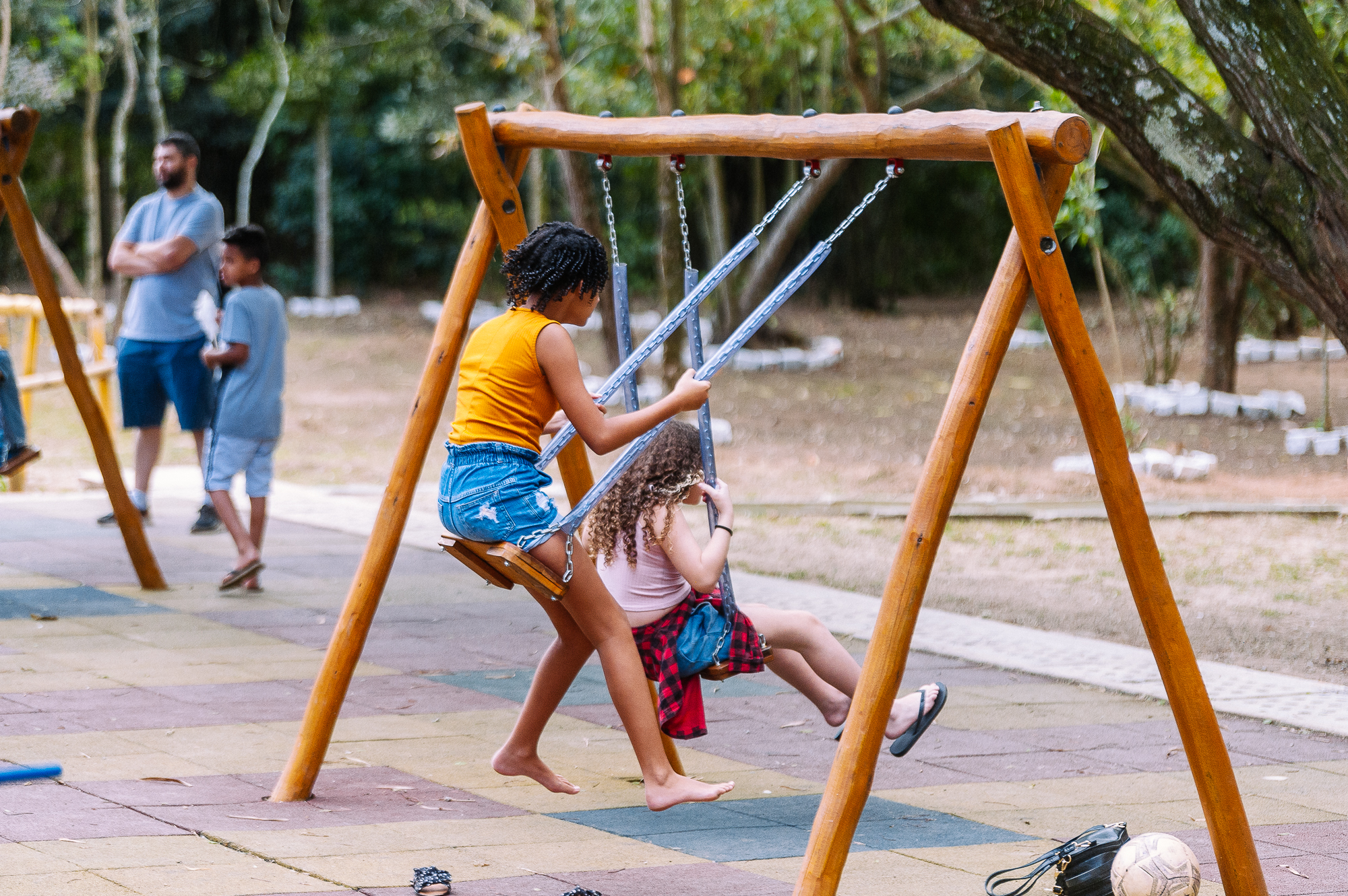 Duas crianças brincam em um balanço de madeira em um parque ao ar livre. O balanço tem estrutura robusta em madeira de tom marrom claro, com correntes metálicas prateadas. A criança à esquerda, com cabelo escuro e cacheado, usa uma regata laranja vibrante e um short jeans azul, e está descalça enquanto empurra o balanço. A criança à direita, sentada, tem cabelo claro e cacheado, veste uma camiseta rosa clara e um short vermelho com estampa xadrez, usando chinelos escuros.  O chão é composto por placas emborrachadas coloridas, em tons de bege, marrom e verde. Ao fundo, há árvores com folhas verdes e um ambiente que mistura áreas de terra e vegetação, criando um cenário natural. Também aparecem, desfocados, um adulto com camiseta cinza e shorts azul-escuro e outra criança com roupas em tons de azul.  No canto inferior direito, há uma bola branca com detalhes em cinza e uma bolsa preta apoiada no chão. A iluminação é natural, com luz suave, sugerindo um momento durante o dia em um ambiente tranquilo e arborizado.