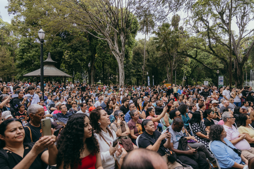 Fotografia mostra uma grande plateia reunida ao ar livre em um parque arborizado. Centenas de pessoas, de diferentes idades, ocupam cadeiras e áreas gramadas enquanto acompanham uma atividade cultural. Muitas seguram celulares para registrar o momento. Ao fundo, árvores de grande porte formam uma extensa cobertura verde, e um coreto aparece entre a vegetação. O ambiente transmite clima de convivência, lazer e participação comunitária, com o público concentrado e atento às apresentações realizadas no parque.