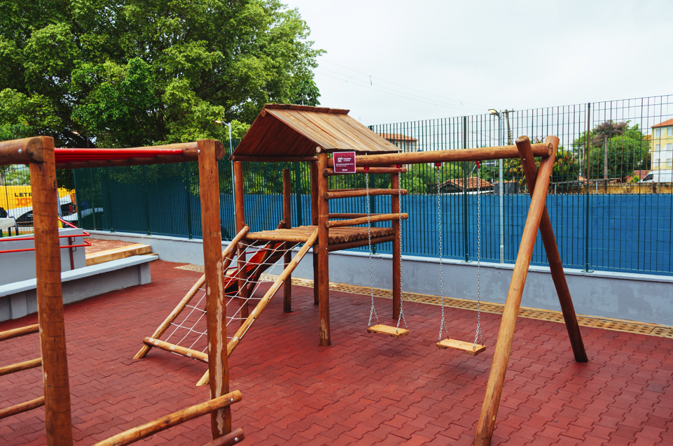 Imagem colorida em plano médio de um parquinho infantil ao ar livre. No centro, há uma estrutura de brinquedo feita de troncos de madeira rústica, composta por uma pequena ponte coberta por um telhado de tábuas, uma rede de escalada de cordas brancas e dois balanços de madeira presos por correntes. O chão é todo revestido com piso intertravado emborrachado na cor vermelha. Ao fundo, uma grade metálica verde delimita o espaço, com árvores frondosas à esquerda e um céu nublado ao fundo. À esquerda, em segundo plano, avista-se uma rampa de concreto cinza com corrimão vermelho.
