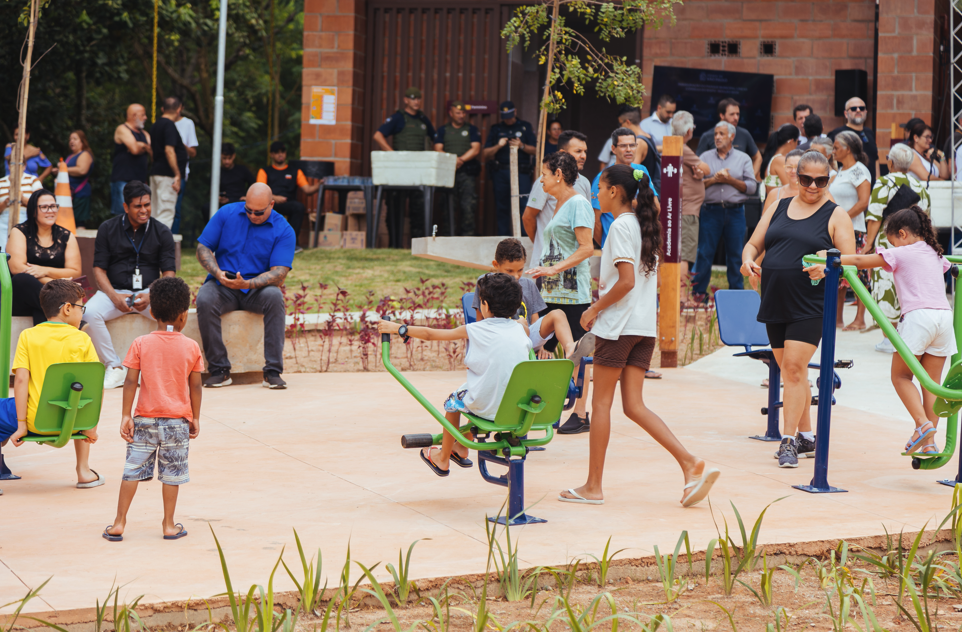 **Descrição resumida:**  A imagem mostra a movimentação de pessoas no Parque Municipal Córrego do Bispo. Em primeiro plano, sobre um piso de concreto claro, várias crianças divertem-se e usam os aparelhos verdes da academia ao ar livre e do parquinho.  Adultos circulam e observam a cena ao redor. Ao fundo, destaca-se a estrutura de tijolos ecológicos aparentes do parque, onde um grupo de pessoas se reúne próximo a um totem de madeira com a inscrição "Academia ao Ar Livre". Três guardas civis acompanham o evento discretamente. O ambiente é cercado por áreas verdes em desenvolvimento, sob a iluminação natural do dia.