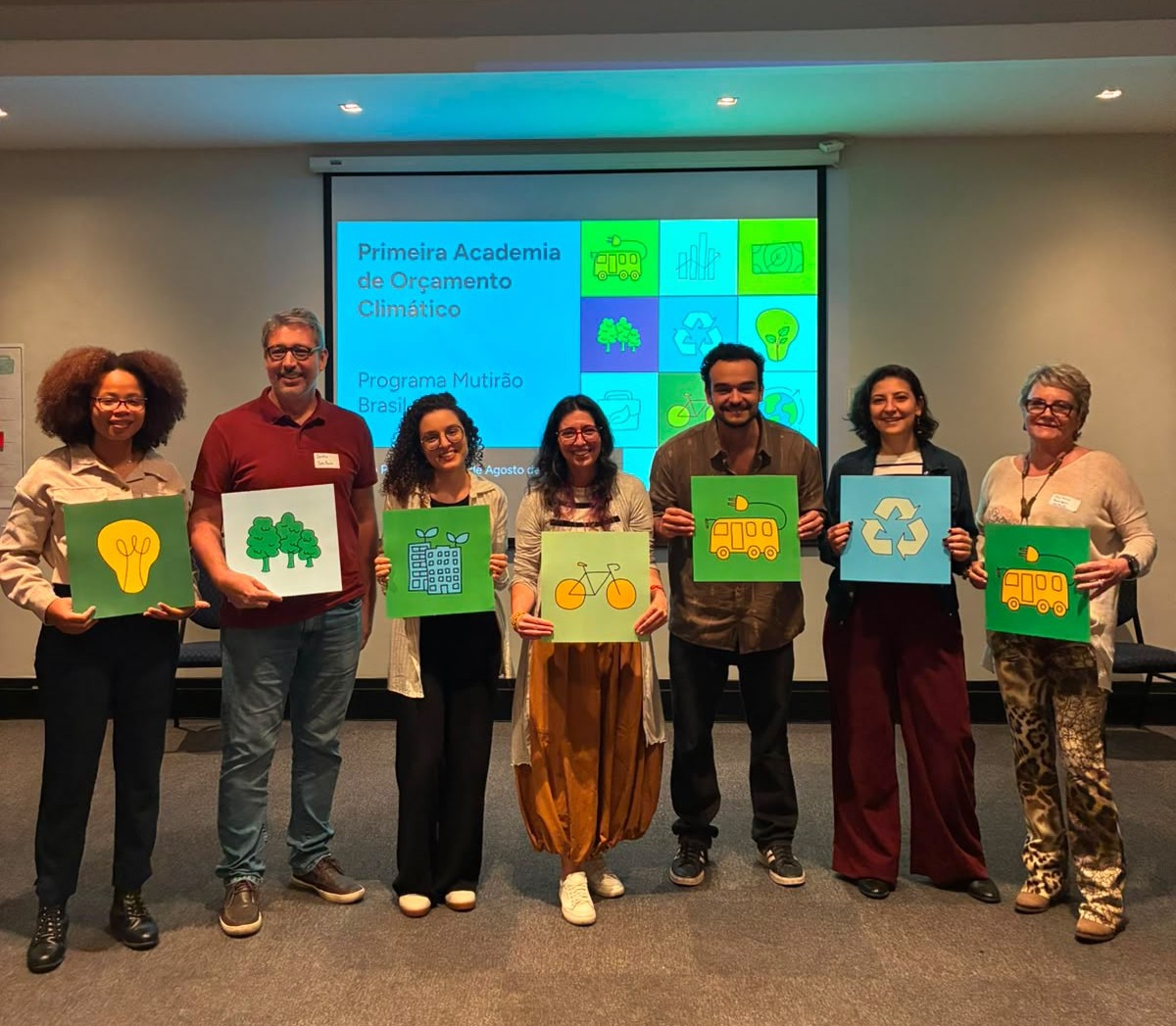 Foto de grupo em uma sala, diante de uma tela de projeção. Sete pessoas estão lado a lado, de pé, segurando placas coloridas com ilustrações relacionadas à sustentabilidade, como árvores, bicicleta, ônibus, reciclagem e lâmpada. Ao fundo, a tela exibe a apresentação “Primeira Academia de Orçamento Climático – Programa Mutirão Brasil”.
