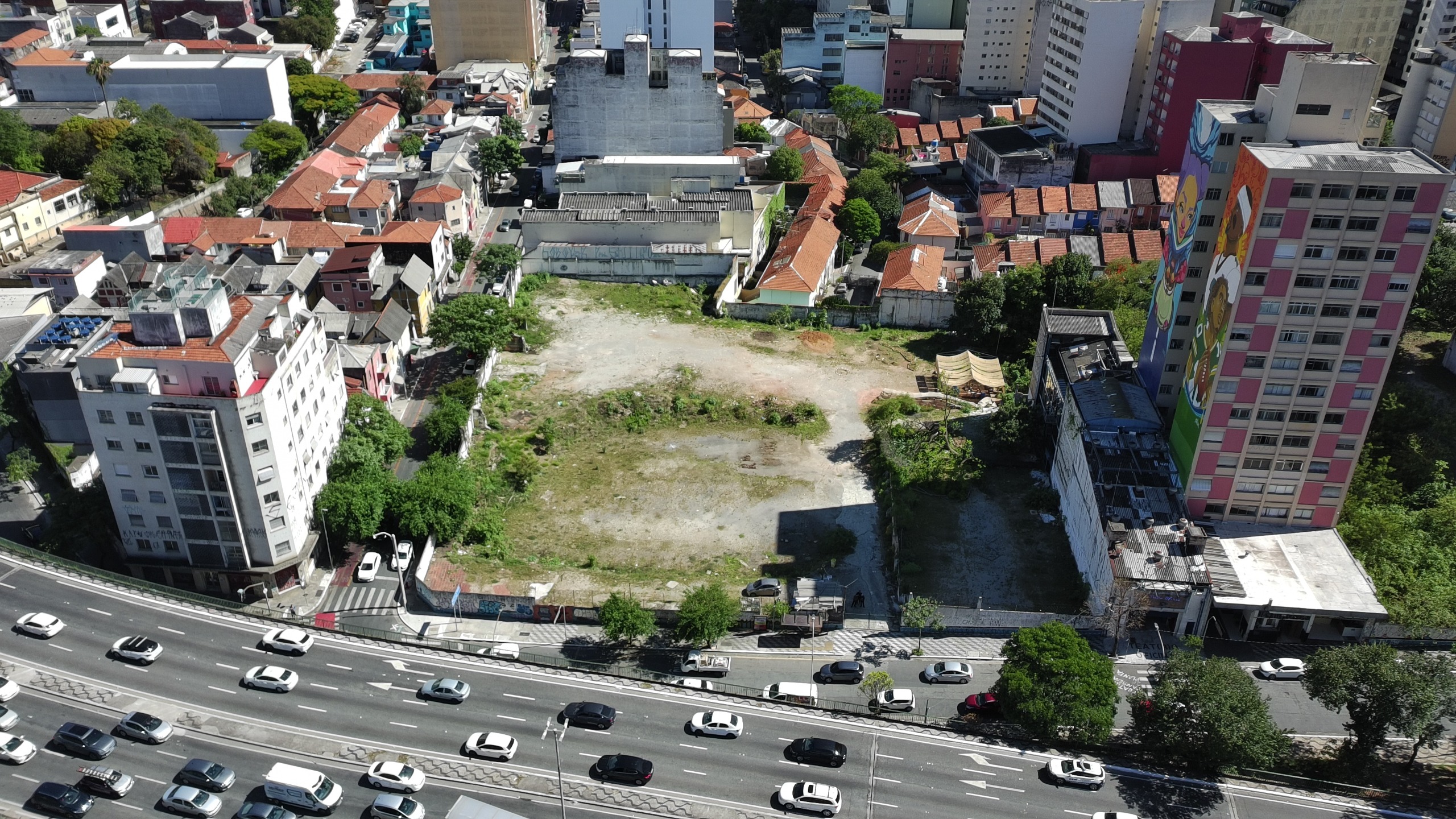 Vista aérea de um terreno baldio retangular no centro de uma área urbana densa. O lote tem solo de terra batida e pouca vegetação, cercado por muros, casas de telhado cerâmico e prédios. À direita, destaca-se um edifício com um grande mural colorido. Na parte inferior, uma avenida larga e movimentada com vários carros. A iluminação é solar e intensa.