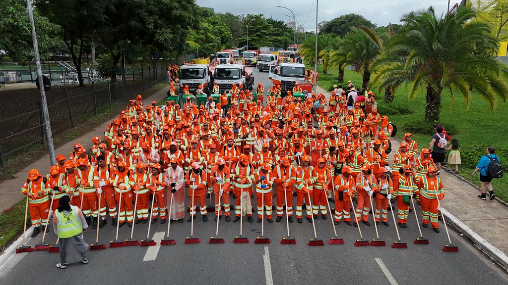 Fotografia colorida em plano aberto e vista superior de uma grande equipe de limpeza urbana posicionada em uma avenida de asfalto escuro. Dezenas de profissionais, homens e mulheres, vestem uniformes na cor laranja vibrante com listras refletivas amarelas e verdes, estando a maioria perfilada e segurando vassouras de cabo longo com cerdas vermelhas voltadas para o chão. Ao fundo, vários caminhões de limpeza brancos estão estacionados em fila atrás do grupo. A via é ladeada por árvores e uma grade metálica à esquerda, e um gramado com palmeiras e alguns pedestres à direita. O céu está nublado e a iluminação é diurna. No canto inferior esquerdo, uma pessoa de costas utiliza um colete amarelo fluorescente sobre uma capa de chuva transparente.