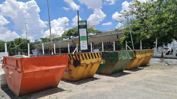 A imagem apresenta uma fileira de quatro grandes caçambas metálicas de entulho alinhadas em um terreno de cascalho claro, sob a luz do sol. Da esquerda para a direita, as caçambas têm as cores laranja, amarela com listras pretas na borda, verde com marcas de ferrugem e amarela com listras pretas. Ao centro, atrás dos recipientes, há um totem informativo vertical com as cores verde, preto e branco, indicando ser um "Ecoponto" da Mooca. No plano de fundo, observa-se a estrutura de um viaduto de concreto, árvores com folhagens verdes e, ao alto, um céu azul intenso com grandes nuvens brancas e volumosas.